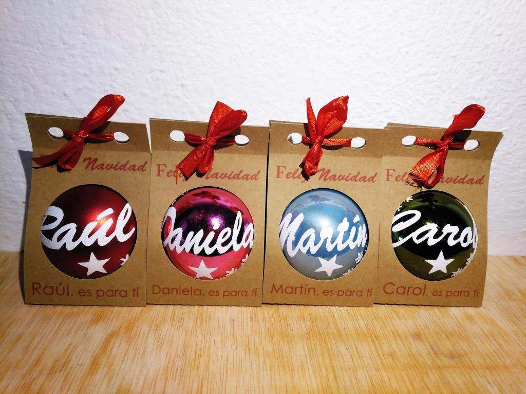 Cuatro bolas de Navidad de colores rojo, fucsia, azul y verde en sus respectivas cajas kraft individuales con lazo rojo y nombres personalizados.