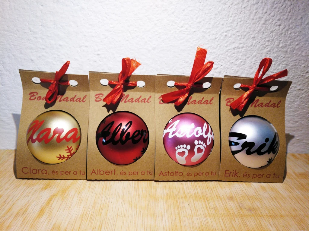 Cuatro bolas de Navidad personalizadas en cajas individuales de cartón kraft con lazo rojo y texto Bon Nadal.