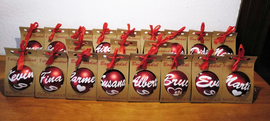 Conjunto de bolas de Navidad rojas personalizadas con nombres en cajas de cartón kraft con lazo rojo dispuestas sobre una superficie de madera.