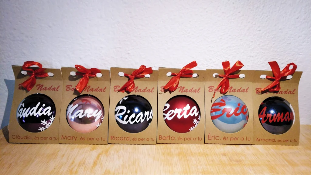 Seis bolas de Navidad de distintos colores presentadas en estuches individuales de cartón kraft con lazo rojo y nombres impresos.