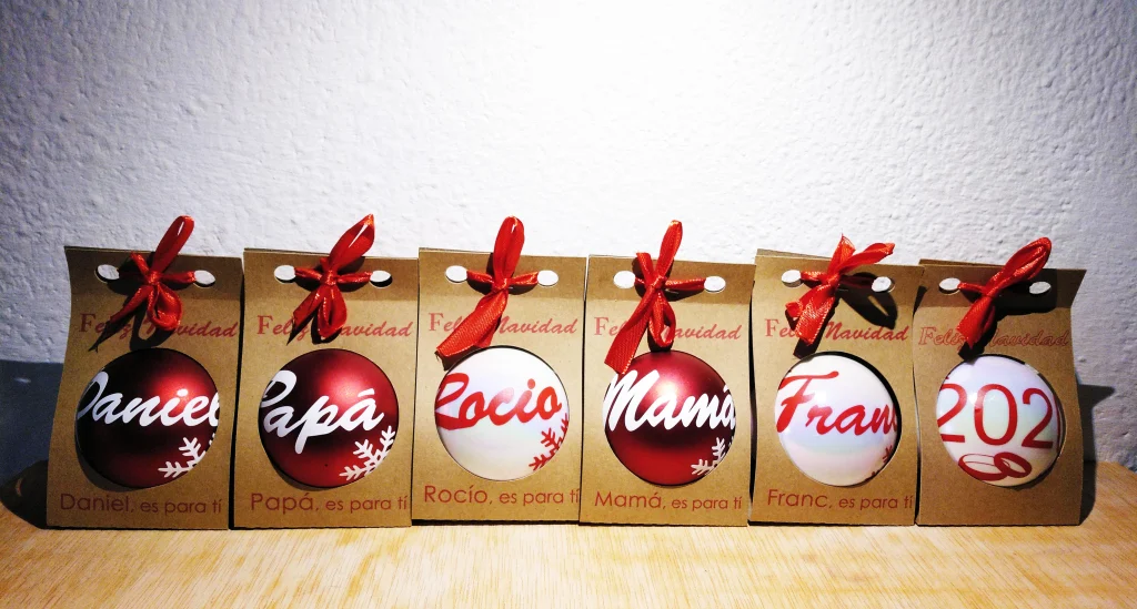 Seis bolas de Navidad personalizadas presentadas en sus respectivas cajas de cartón kraft con lazo rojo.