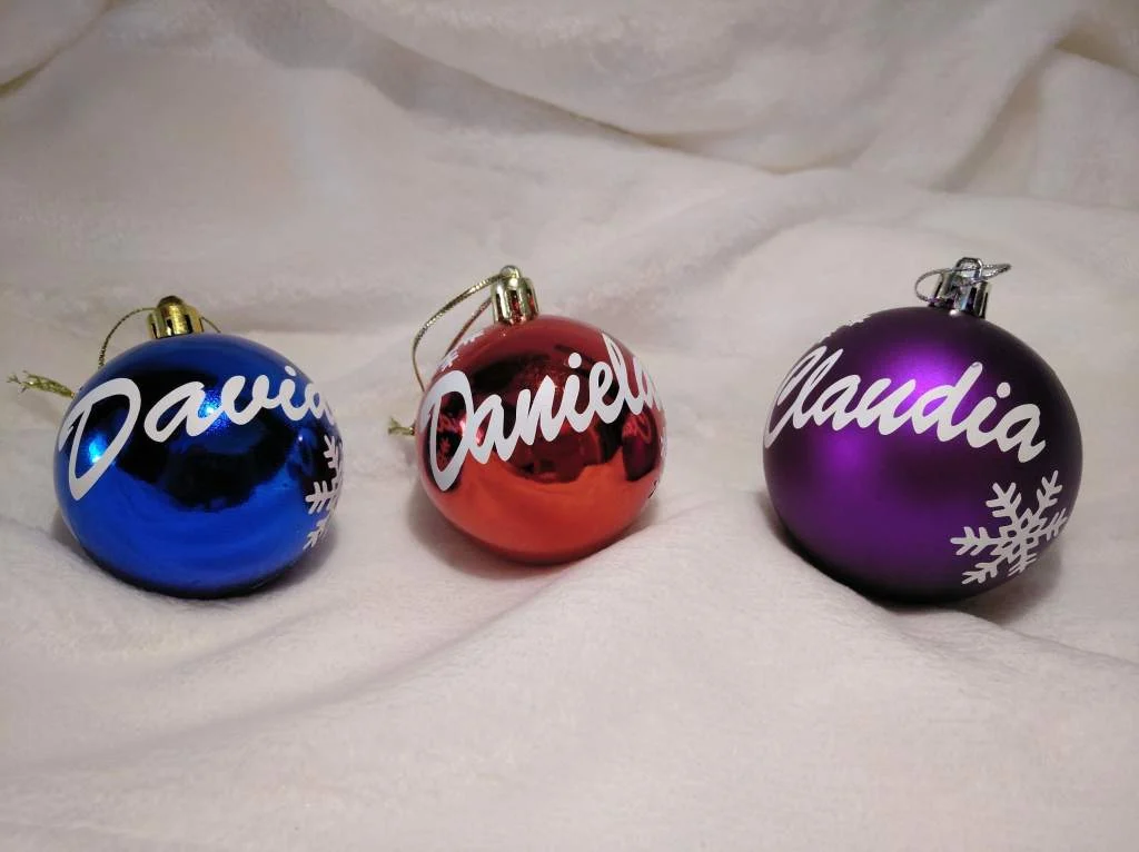 Tres bolas de Navidad personalizadas en colores azul brillante, rojo brillante y morado mate, cada una con un nombre diferente escrito en blanco.