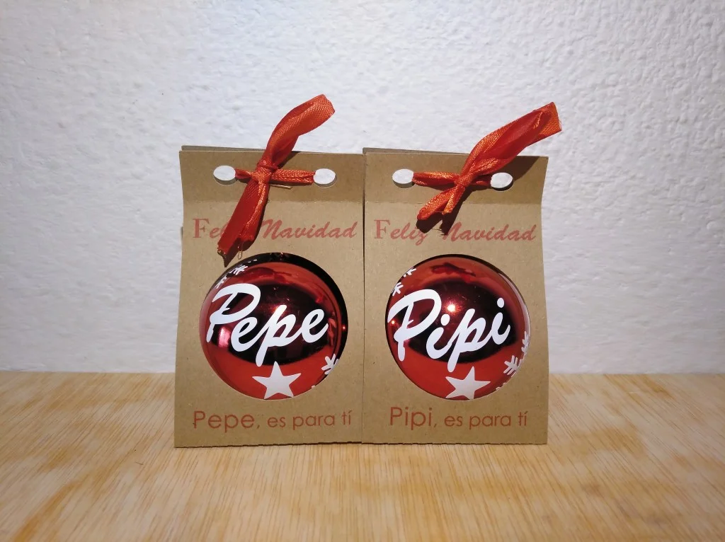 Dos bolas de Navidad rojas y brillantes personalizadas con los nombres Pepe y Pipi en blanco, presentadas en cajitas de cartón kraft con lazo rojo.