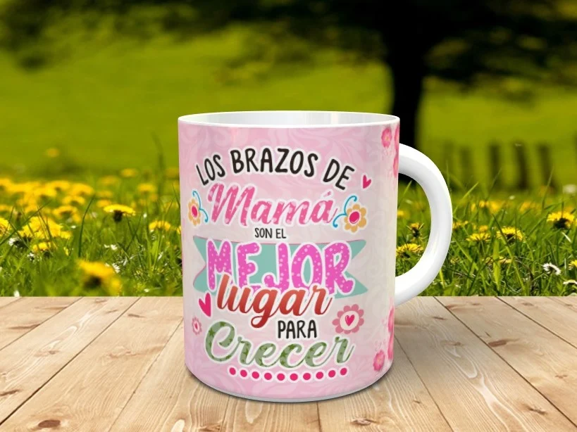 Taza blanca personalizable con el mensaje Los brazos de mamá son el mejor lugar para crecer sobre fondo rosado, apoyada en madera.