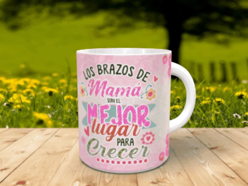 Taza blanca personalizable con un diseño frontal dedicado a los abrazos de mamá sobre un fondo rosa con flores y corazones, apoyada en una superficie de listones de madera ante un paisaje natural difuminado.
