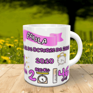 Taza blanca personalizable con los datos de nacimiento de la niña Paula en tonos rosas, apoyada sobre una superficie de madera ante un fondo natural desenfocado.