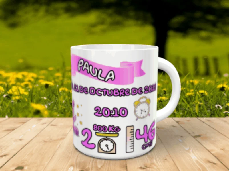 Taza blanca personalizable con los datos de nacimiento de la niña Paula en tonos rosas, apoyada sobre una superficie de madera ante un fondo natural desenfocado.