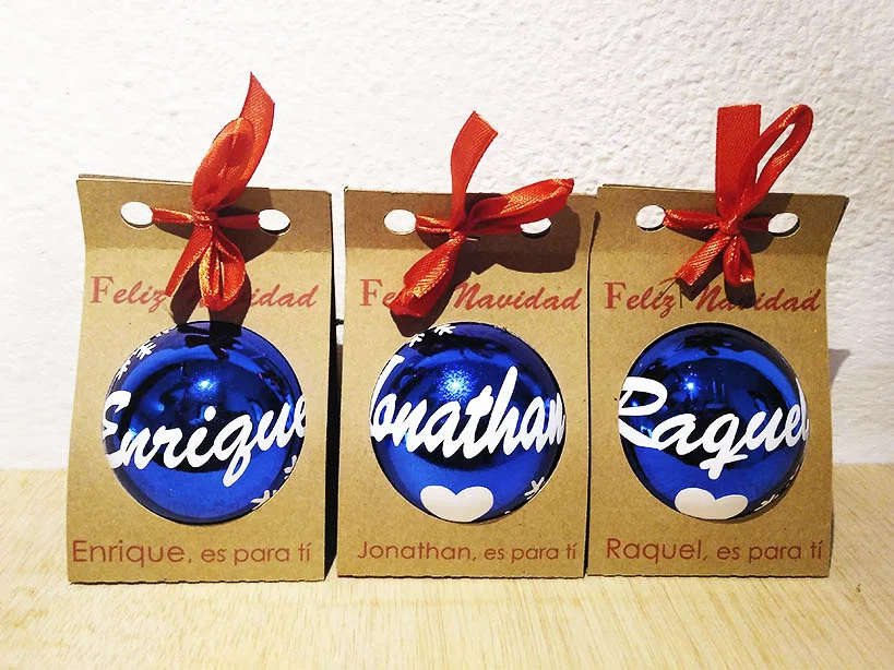 Tres bolas de Navidad azules brillantes con nombres impresos en blanco, presentadas en estuches individuales de cartón kraft con ventana circular, lazo rojo y felicitación.