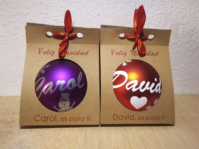 Dos bolas de Navidad personalizadas en estuches individuales de cartón kraft con ventana circular y lazo rojo. Una bola es morada con un muñeco de nieve y el nombre Carol; la otra es roja con un corazón y el nombre David.