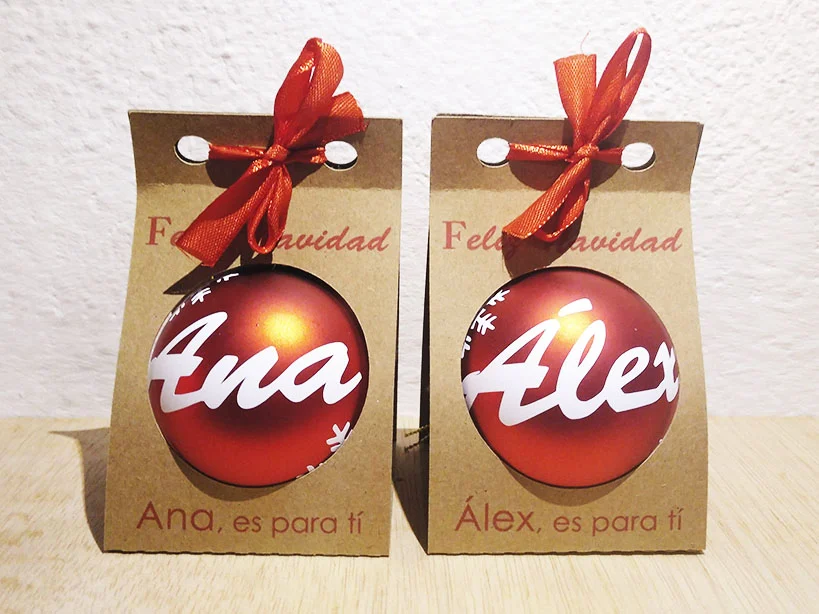 Dos bolas de Navidad rojas personalizadas con los nombres Ana y Alex, presentadas en estuches individuales de cartón kraft con lazo rojo.