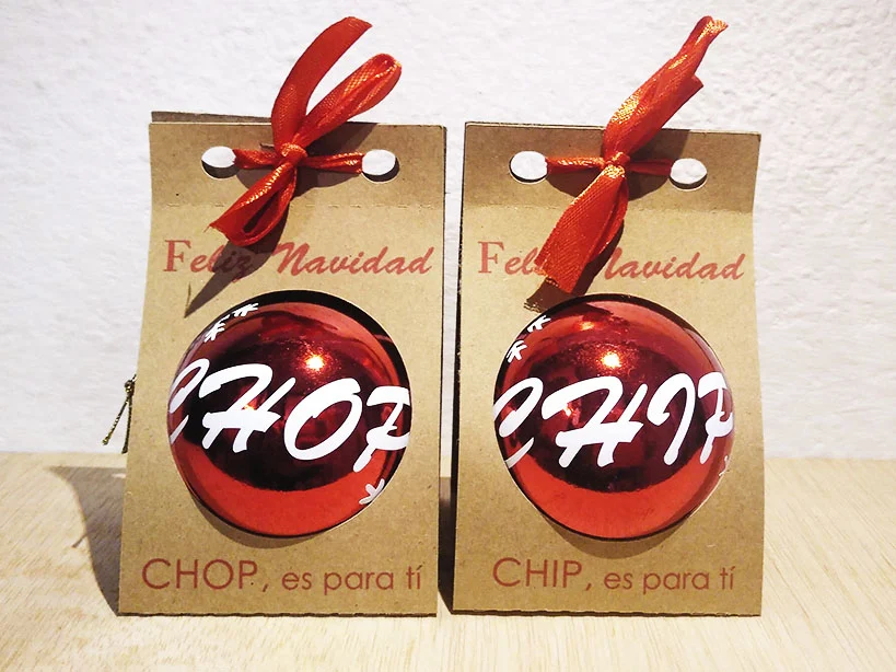 Dos bolas de Navidad rojas brillantes con los nombres CHIP y CHOP impresos en blanco, presentadas en estuches individuales de cartón kraft con ventana circular y lazo rojo.