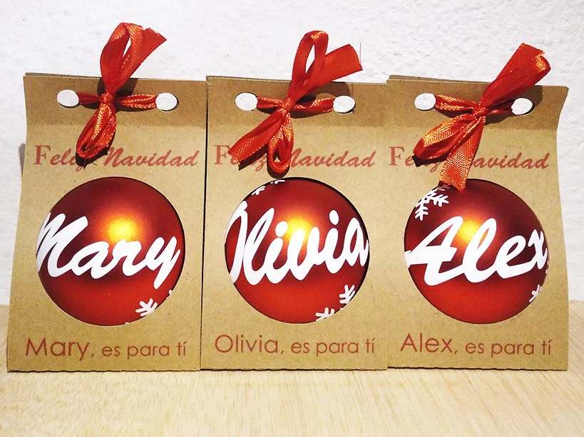 Tres bolas de Navidad rojas personalizadas con nombres en blanco y presentadas en estuches de cartón kraft con lazo rojo.