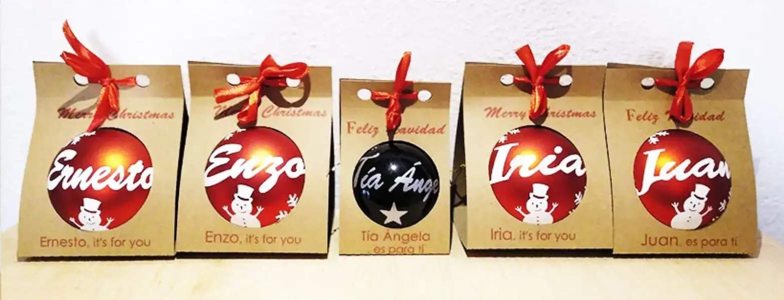 Cinco cajas kraft con ventana circular que muestran bolas de Navidad personalizadas con nombres y motivos festivos, atadas con un lazo rojo.