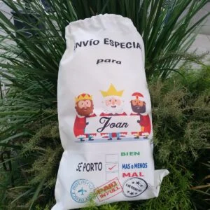 Saco blanco de tela personalizado con la ilustración de los Reyes Magos y el nombre Joan impresos en el frontal, colocado sobre plantas en exterior.