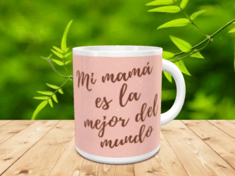 Taza blanca de cerámica personalizada con fondo rosa y texto Mi mamá es la mejor del mundo, sobre superficie de madera con vegetación de fondo.