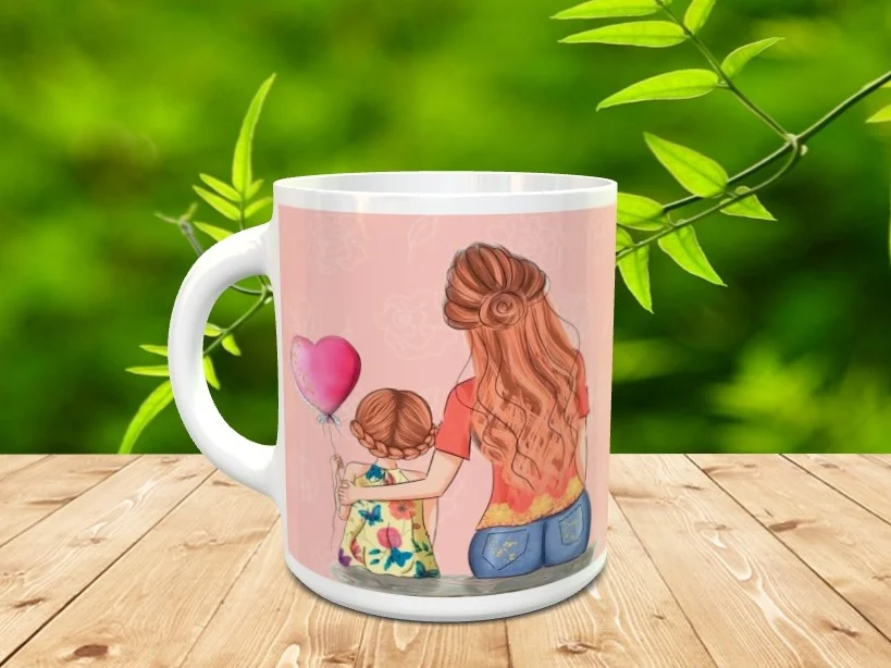 Taza blanca de cerámica personalizada con una tierna ilustración de una madre y su hija abrazadas sosteniendo un globo con forma de corazón. La pieza descansa sobre una superficie de madera con vegetación al fondo.