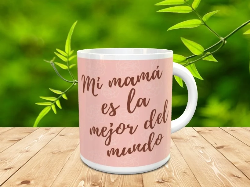 Taza blanca de cerámica personalizada con el texto Mi mamá es la mejor del mundo sobre un fondo rosa con motivos florales, apoyada en una superficie de madera.