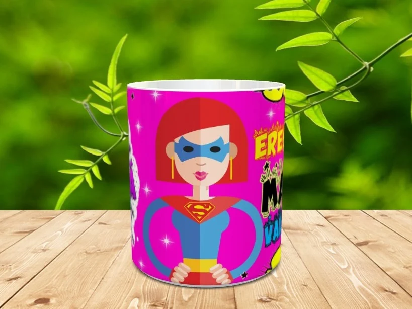 Taza de cerámica blanca personalizada con la ilustración de una mamá superheroína sobre un fondo fucsia con el texto Eres una mamá valiente, apoyada sobre una superficie de madera.