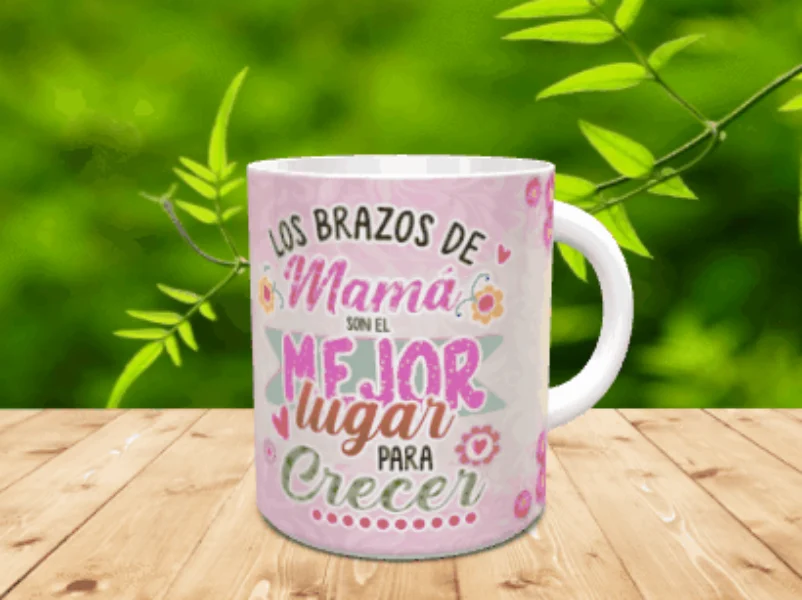 Taza de cerámica blanca con asa, decorada con un diseño en tonos rosas y texto emotivo dedicado a las madres. La pieza reposa sobre una superficie de madera con vegetación de fondo.