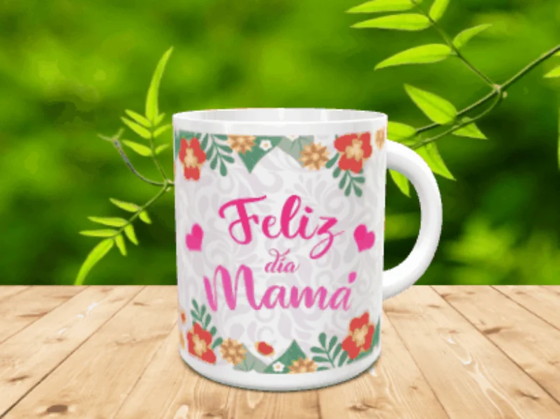 Taza blanca de cerámica personalizada con el texto Feliz día Mamá en tonos rosas, rodeada de detalles florales y corazones, colocada sobre una superficie de madera con fondo verde difuminado.