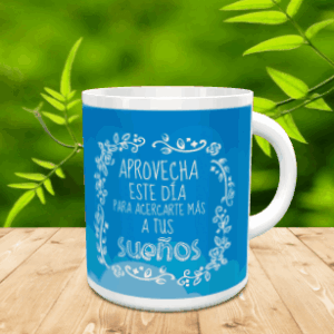 Taza de cerámica blanca con el mensaje APROVECHA ESTE DÍA PARA ACERCARTE MÁS A TUS SUEÑOS dentro de un marco floral sobre fondo azul, apoyada sobre una superficie de madera.
