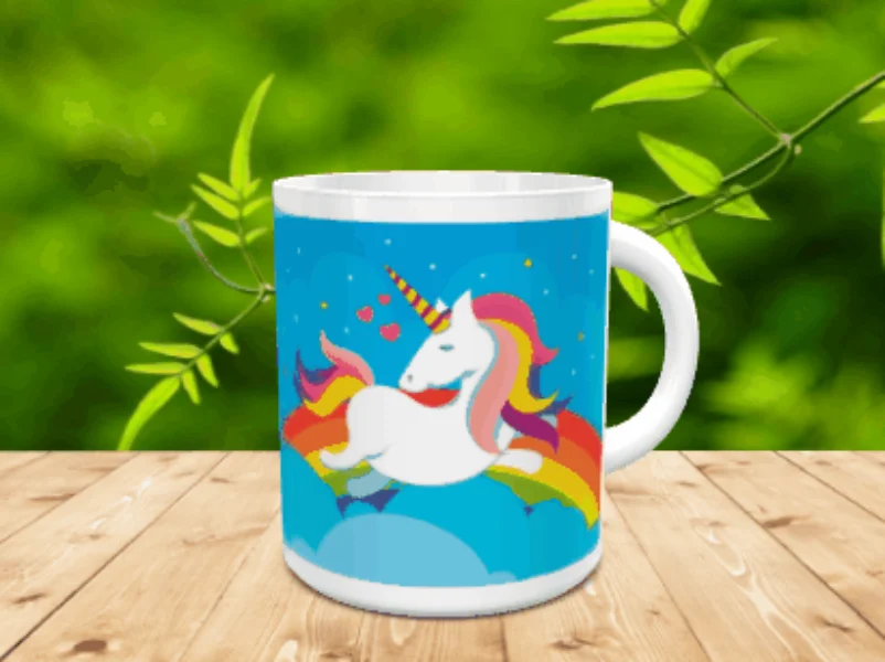 Taza de cerámica blanca con un diseño infantil de unicornio sobre un arco iris y fondo azul de nubes y estrellas. La pieza se sitúa sobre una superficie de madera.
