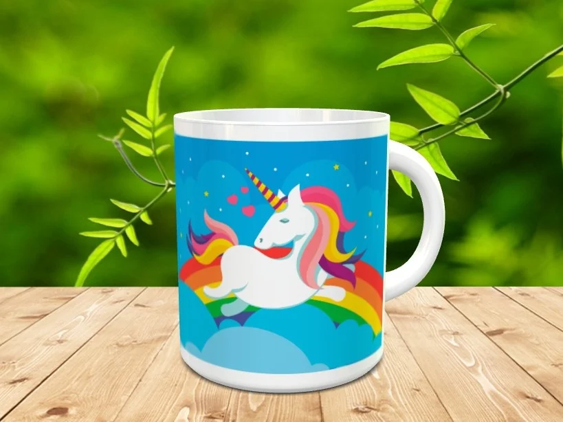 Taza de cerámica blanca con diseño de un unicornio sobre un arco iris y fondo azul, apoyada sobre una superficie de madera.
