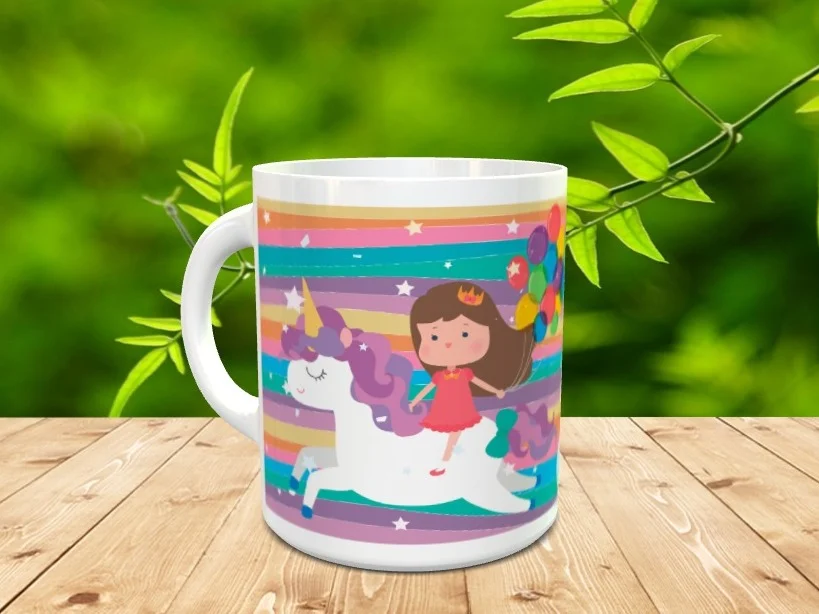Taza de cerámica blanca decorada con la ilustración de una niña sobre un unicornio y globos coloridos sobre fondo de rayas, apoyada en superficie de madera.