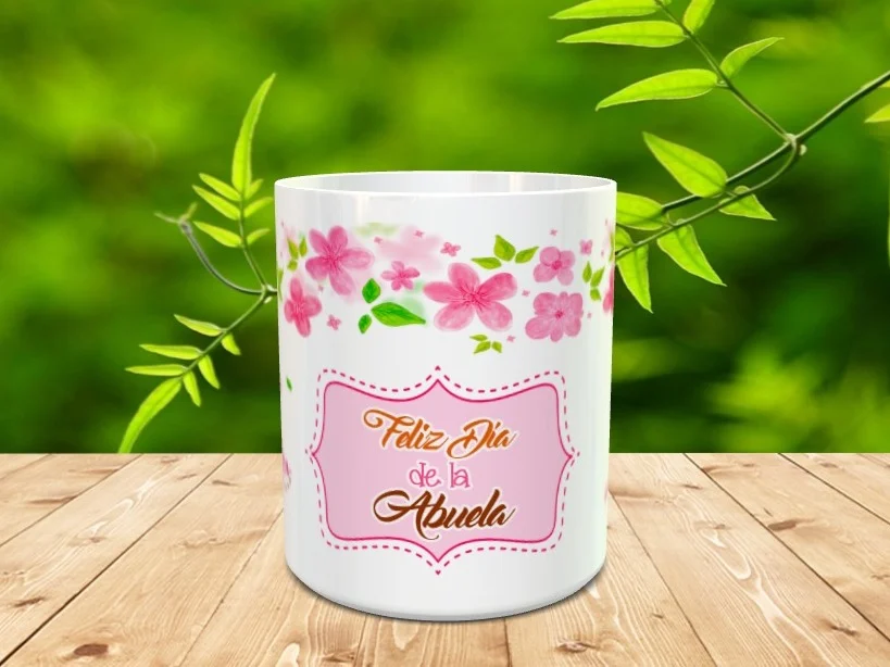 Taza blanca de cerámica decorada con flores rosas y un marco festoneado con el texto Feliz Día de la Abuela sobre una superficie de madera.