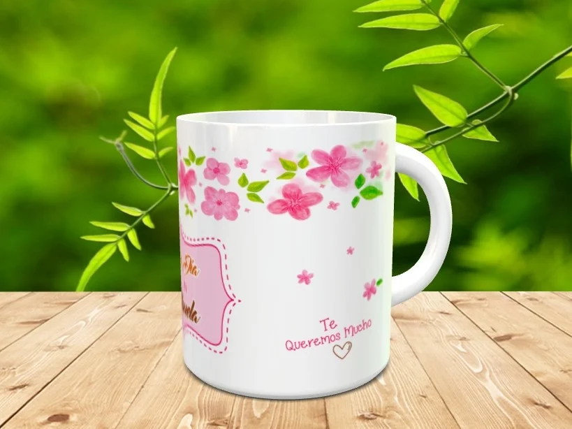 Taza blanca de cerámica para abuelos decorada con flores rosas en la parte superior y un mensaje con marco rosa, situada sobre una superficie de listones de madera.