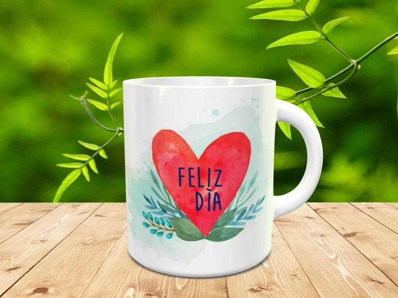 Taza blanca de cerámica decorada con un gran corazón rojo y la frase Feliz Día sobre un fondo de ramas verdes y acuarela, colocada sobre una superficie de madera.