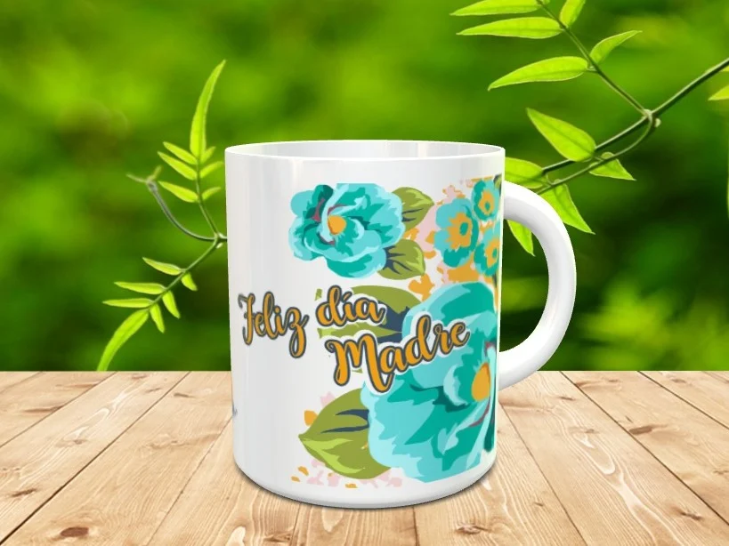 Taza blanca de cerámica decorada con flores turquesas y el mensaje Feliz día Madre sobre una superficie de madera y fondo verde.