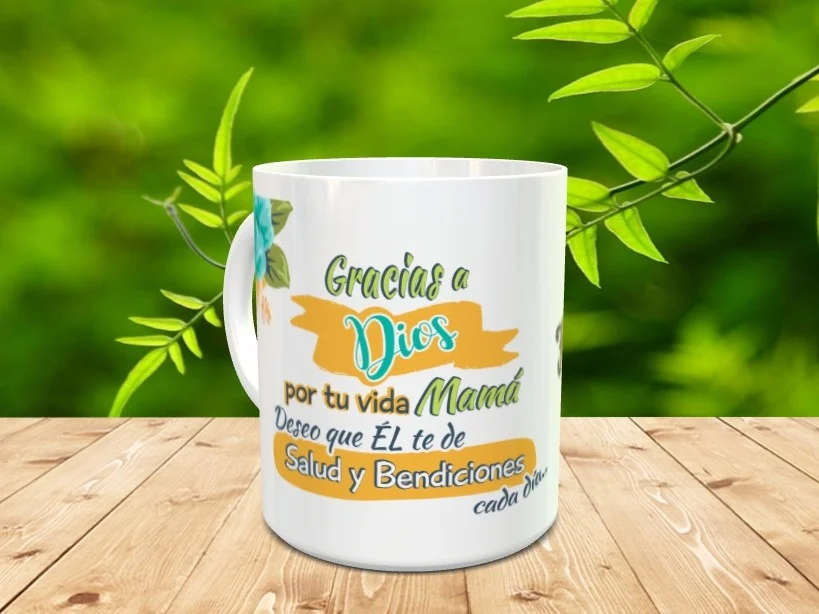 Taza blanca de cerámica decorada con un mensaje de agradecimiento en tonos verdes, azules y amarillos, situada sobre una superficie de madera con fondo vegetal.