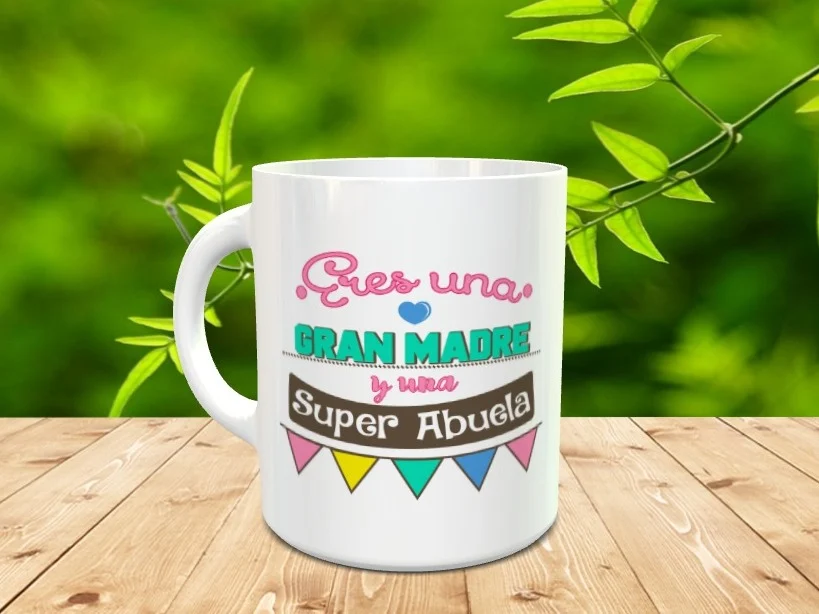 Taza blanca de cerámica decorada con el mensaje Eres una gran madre y una super abuela junto a banderines de colores sobre una superficie de madera.
