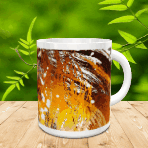 Taza blanca de cerámica decorada con un diseño abstracto en tonos marrones y amarillos, colocada sobre una mesa de madera con fondo verde difuminado.