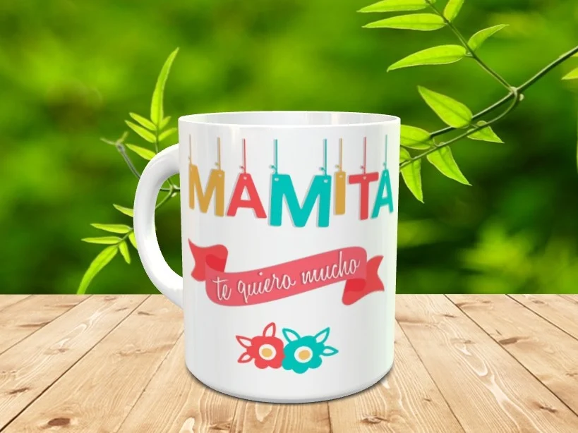 Taza blanca de cerámica para el Día de la Madre decorada con la palabra Mamita colgando de etiquetas de colores y una cinta roja con la frase te quiero mucho. Se sitúa sobre una superficie de madera.