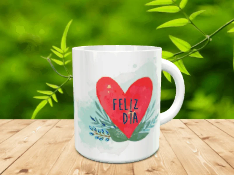 Taza blanca de cerámica en movimiento decorada con un gran corazón rojo y la frase Feliz Día sobre fondo acuarelado y ramas verdes, situada sobre una superficie de madera.