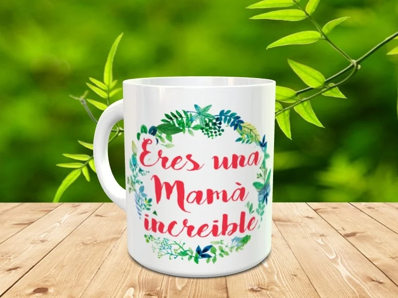 Taza blanca de cerámica decorada con una corona de hojas verdes y azules y la frase Eres una Mamá increíble en tonos rojos. Se presenta sobre una superficie de madera con fondo natural desenfocado.