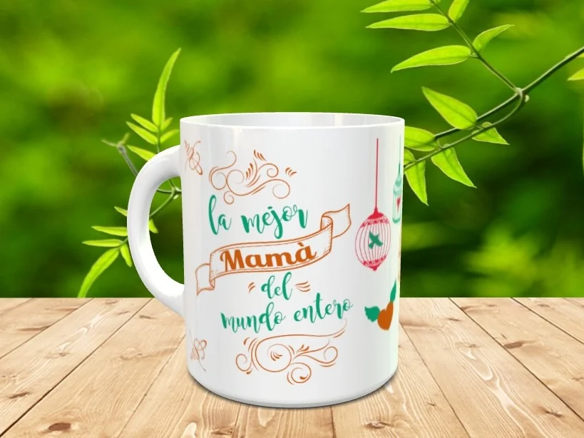 Taza blanca de cerámica para el día de la madre con texto la mejor Mamá del mundo entero y elementos decorativos colgantes sobre superficie de madera. Ref. mama-24xx