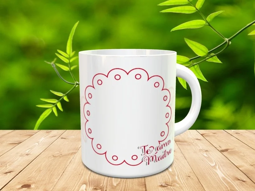 Taza blanca de cerámica con un marco circular festoneado rojo y el texto Te amo Madre, sobre una superficie de madera y fondo vegetal difuminado.
