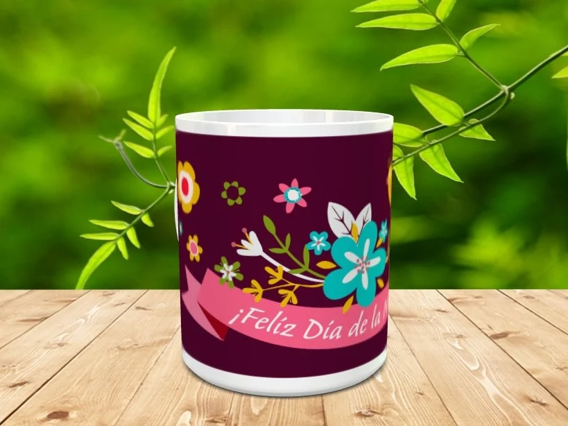 Taza blanca de cerámica decorada con fondo morado, flores coloridas y una cinta rosa con el texto ¡Feliz Día de la Madre sobre una superficie de madera.