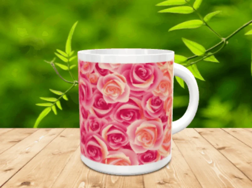 Taza blanca de cerámica decorada con un diseño integral de rosas en tonos rosados y anaranjados, situada sobre una superficie de madera frente a un fondo vegetal desenfocado.