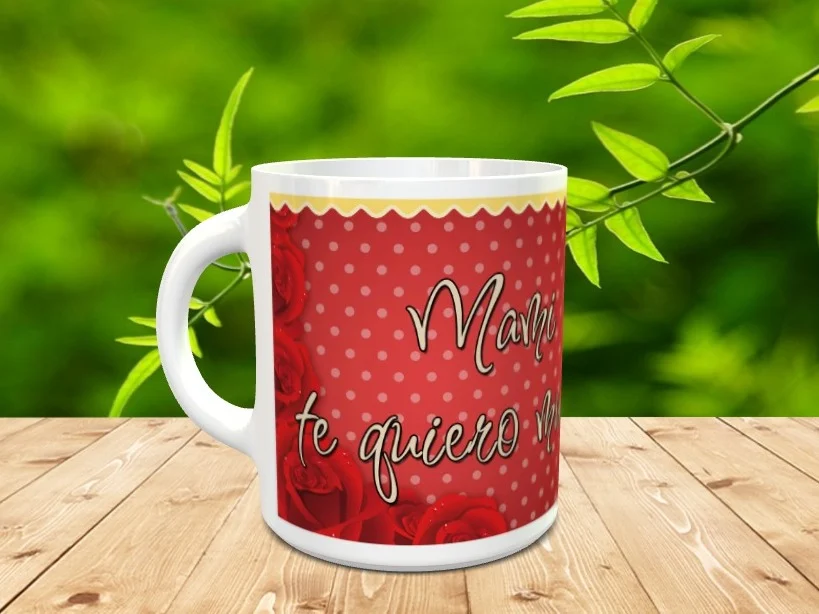 Taza blanca de cerámica con diseño en tonos rojos de rosas y lunares, con la frase Mami te quiero mucho, sobre superficie de madera.
