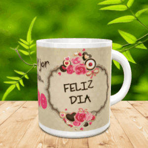 Taza blanca de ceramica con un diseño centrado que muestra un marco decorado con rosas y la frase Feliz Día sobre una superficie de madera y fondo natural difuminado.