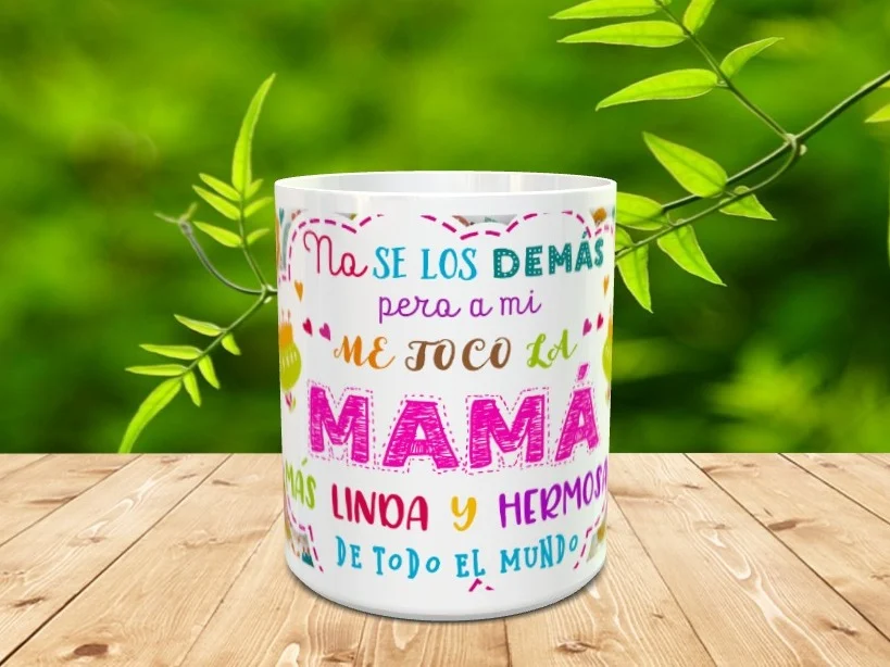 taza blanca de cerámica con un diseño colorido dedicado a las madres, colocada sobre una superficie de madera con fondo de vegetación desenfocada. Ref. mama-54x