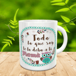 Taza blanca de cerámica con un diseño dedicado a la madre que incluye un marco turquesa, la frase Todo lo que soy te lo debo a ti Mamá y un simpático búho, apoyada sobre una superficie de madera con fondo natural desenfocado.