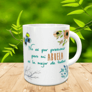 Taza blanca de cerámica decorada con la frase No es por presumir pero mi abuela es la mejor de todas, acompañada de mariposas y flores sobre una superficie de madera.