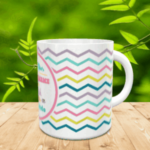 Taza blanca de cerámica con cenefas en zigzag de colores y un círculo central con texto, apoyada sobre una superficie de madera. Ref. mama-9x