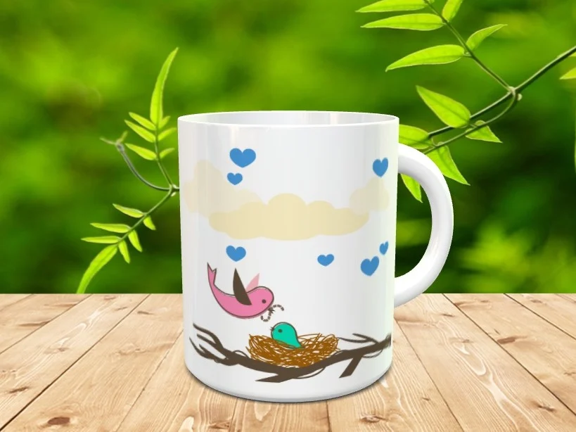 Taza blanca de cerámica decorada con un nido de pájaros, nubes y corazones azules sobre una superficie de madera y fondo vegetal desenfocado.