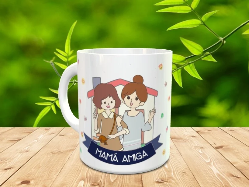 Taza de cerámica blanca con ilustración de dos mujeres y el texto Mamá Amiga, apoyada sobre madera con fondo natural difuminado.